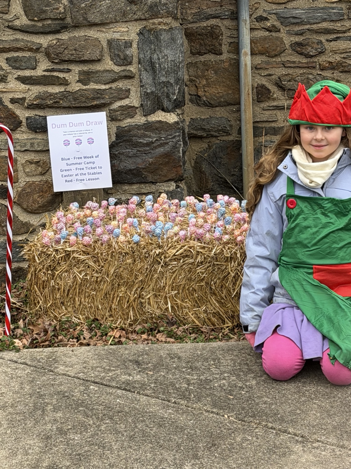 A young festive helper in an elf hat beside a colorful lollipop display at the stone barn
