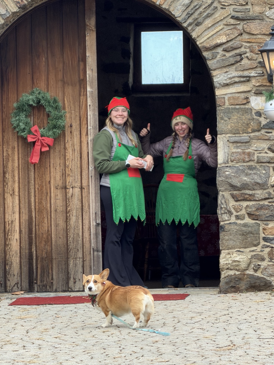 Two festive helpers in elf hats welcome visitors at the stone barn doorway, with a corgi in front