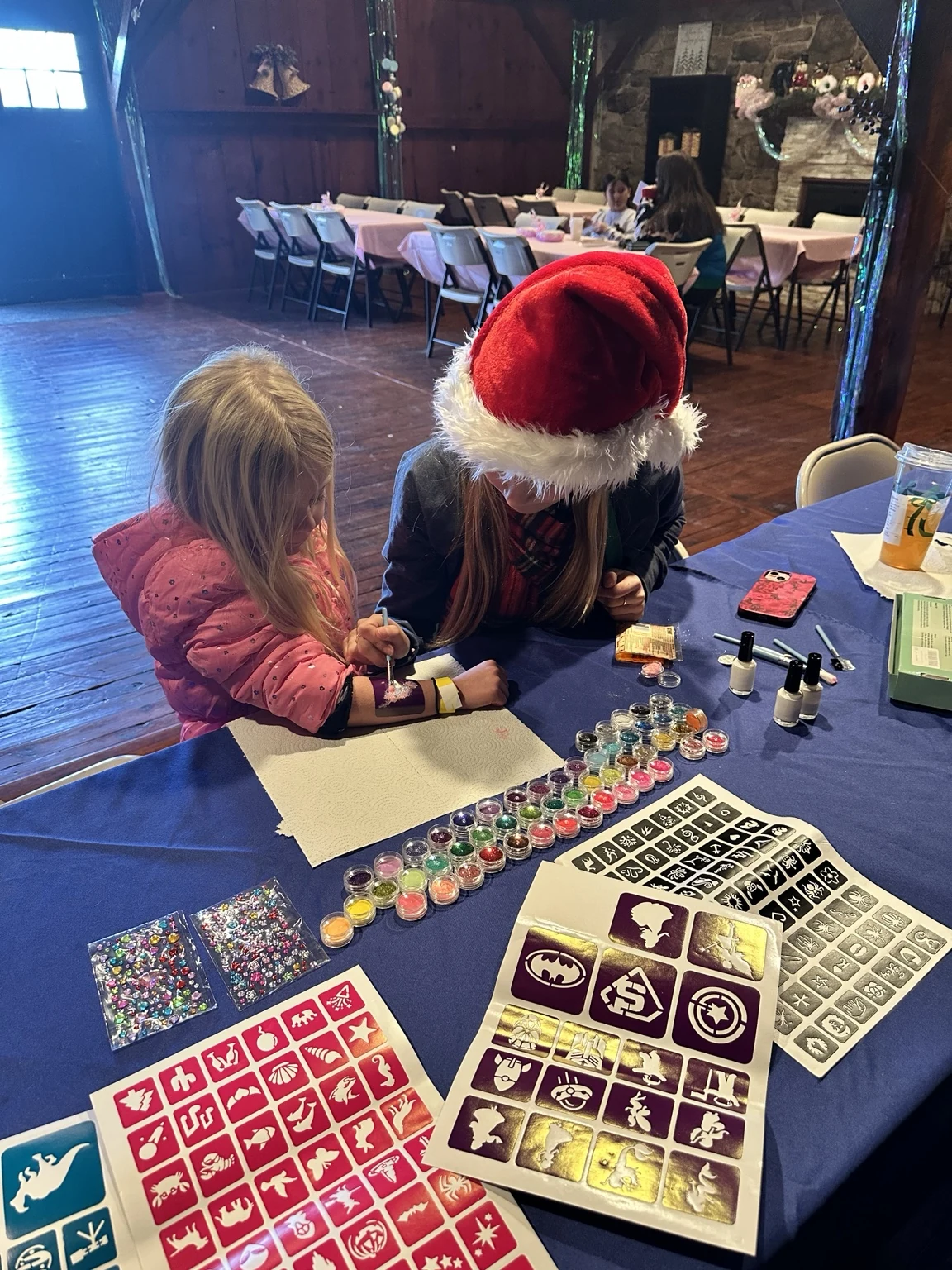 A helper wearing a Santa hat carefully applying a glitter design to a child’s arm at the activity table