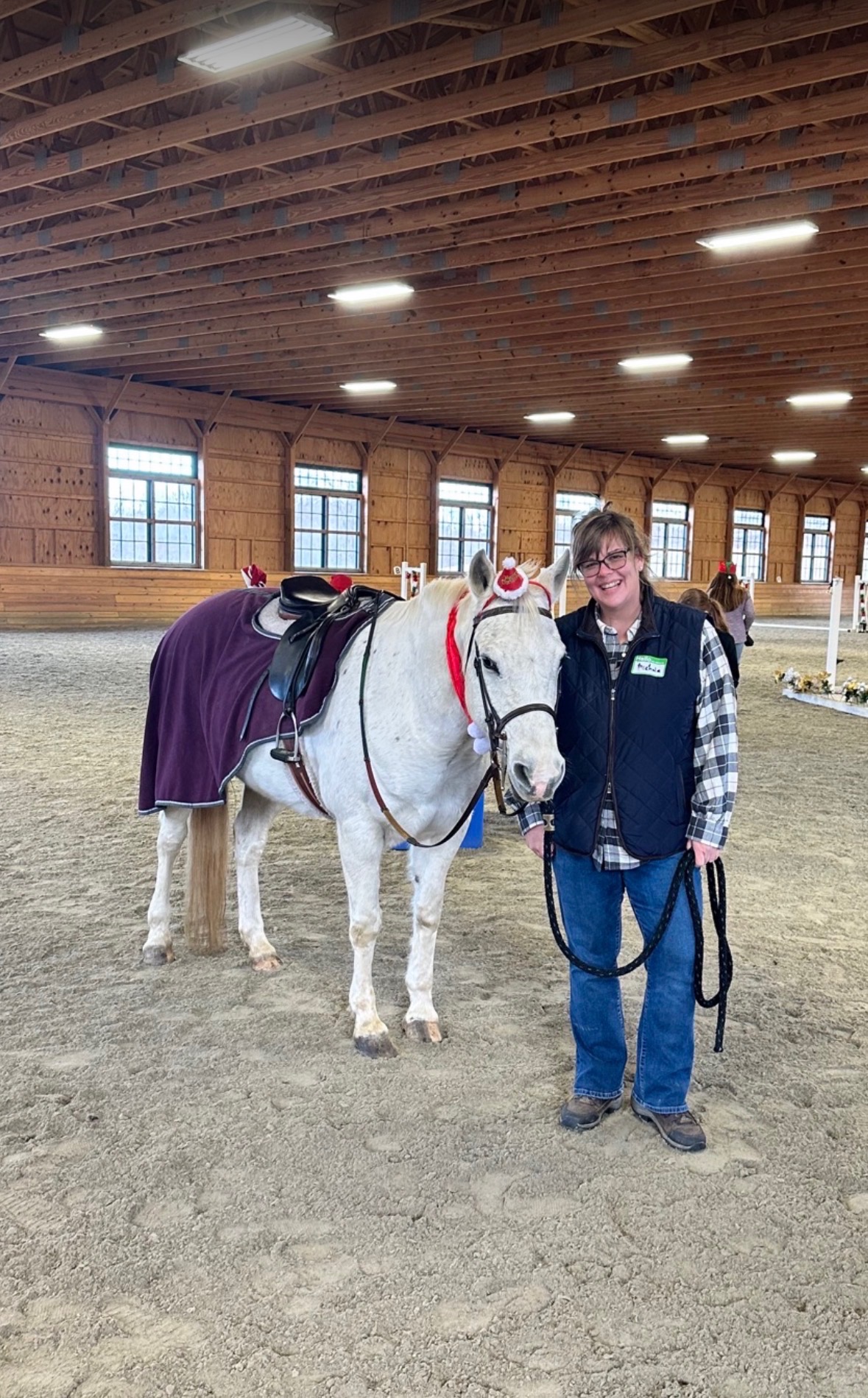A smiling handler with a white pony wearing a festive Santa hat in the indoor arena