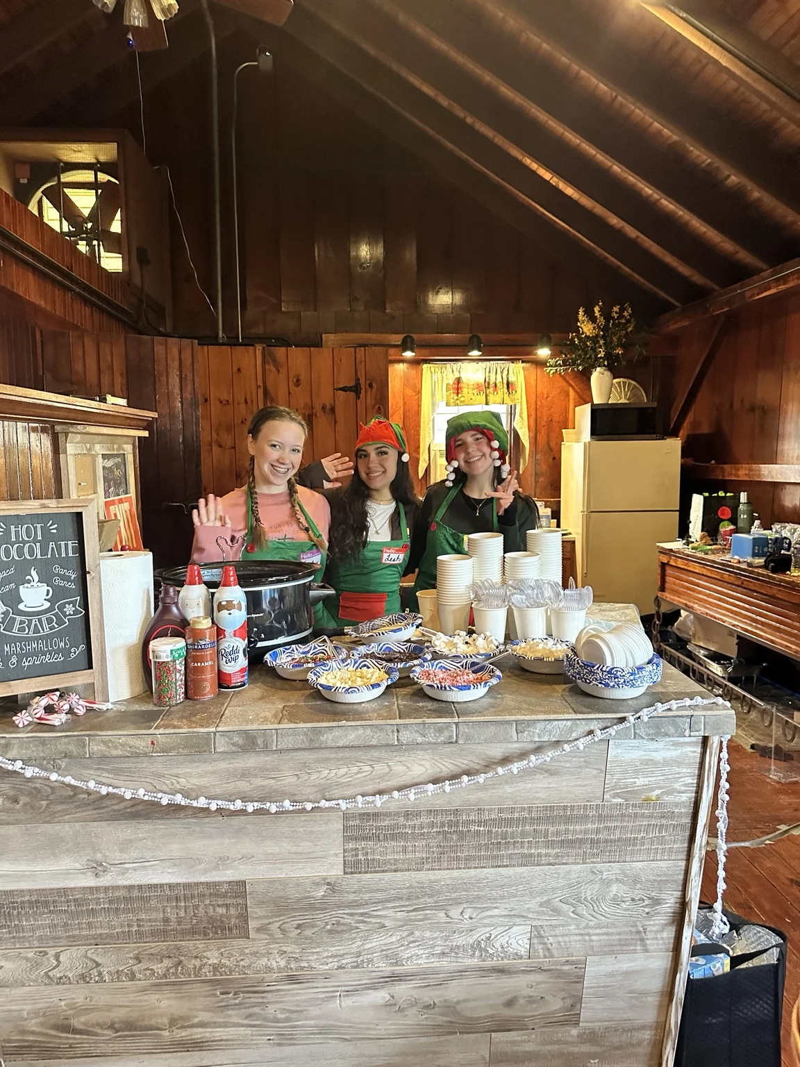 Three smiling holiday helpers behind the farm’s hot chocolate bar with cups, whipped cream and toppings
